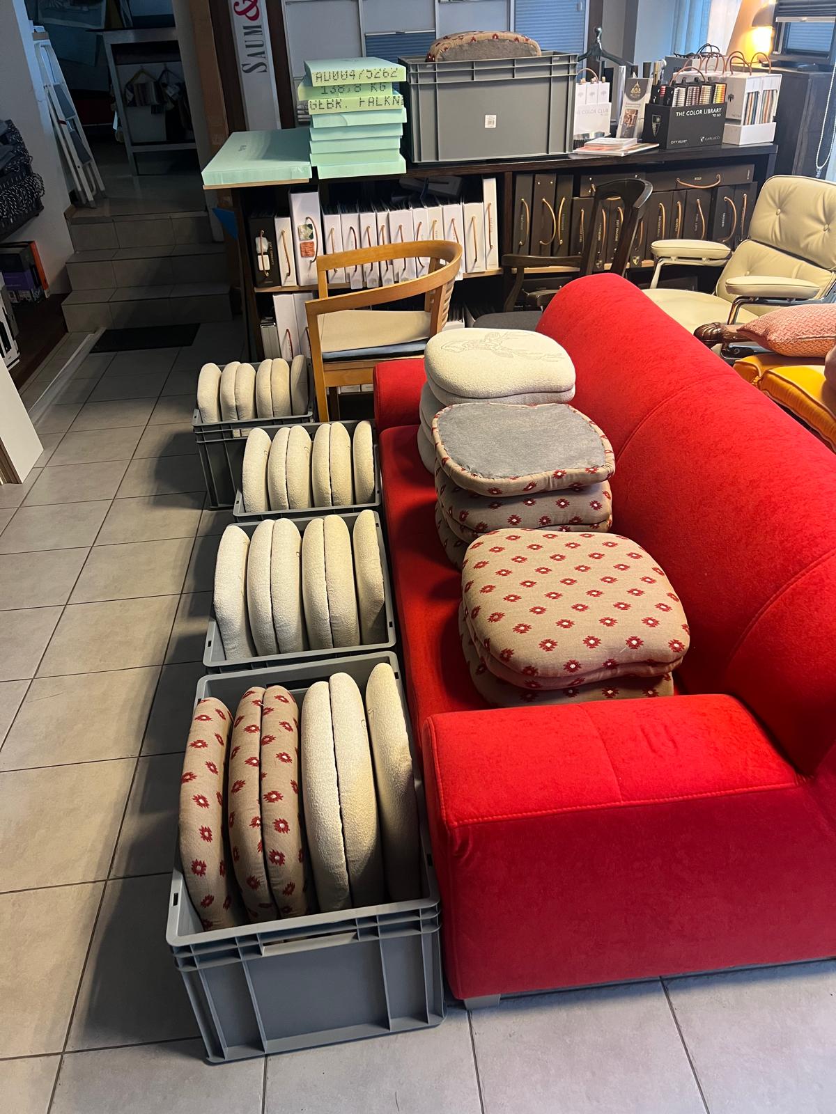 Gefertigte Sitzkissen in Bouclé und gemustertem Stoff, gestapelt neben einem roten Sofa.